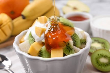 Pouring honey onto delicious fruit salad in bowl on table, closeup