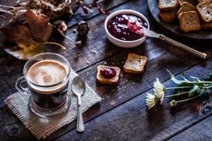 High angle view of a glass coffee cup on rustic wooden table. Small toasted bread slices with marmalade on top are beside the coffee cup.